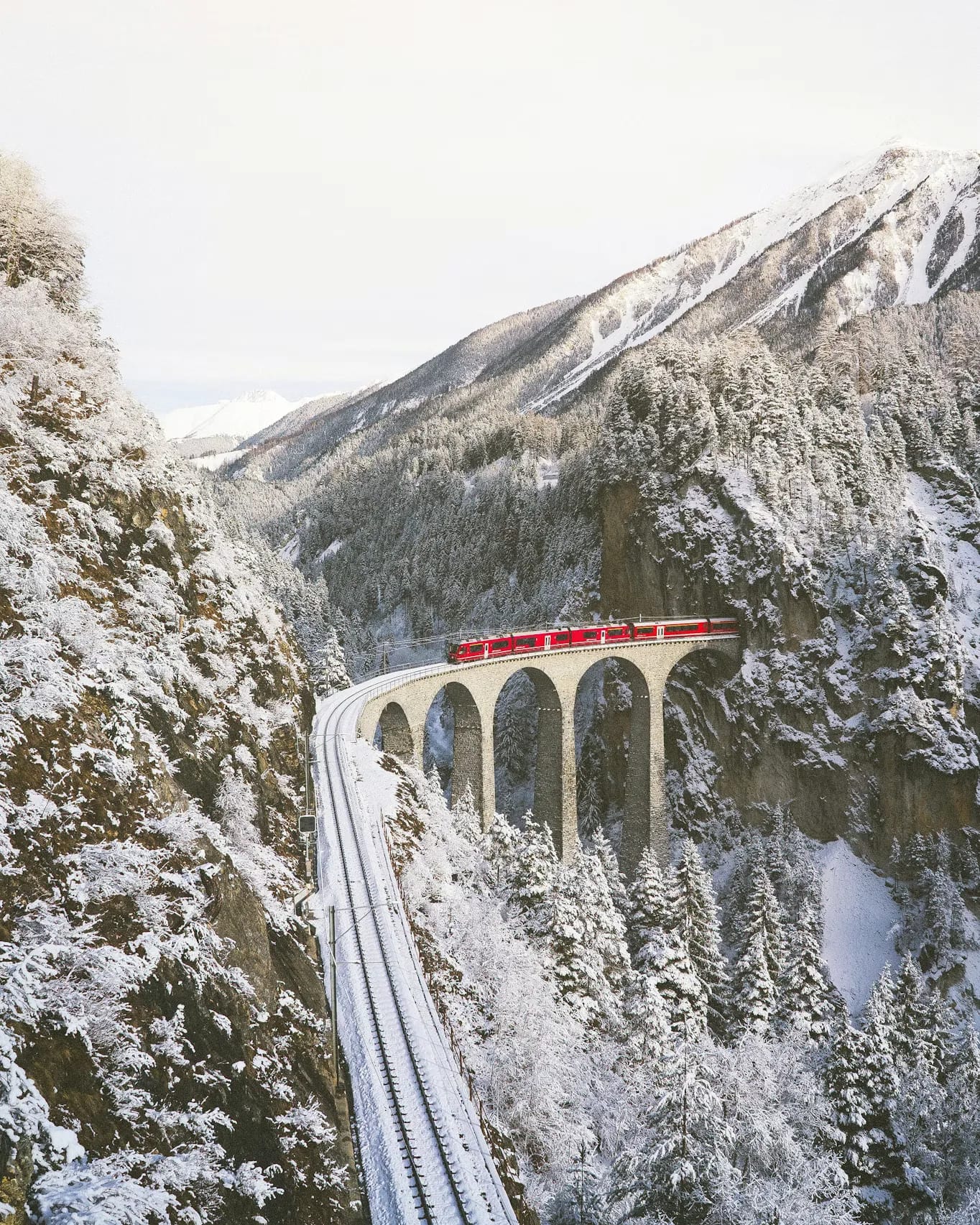 A red train crossing a stone viaduct in a snowy Alpine valley